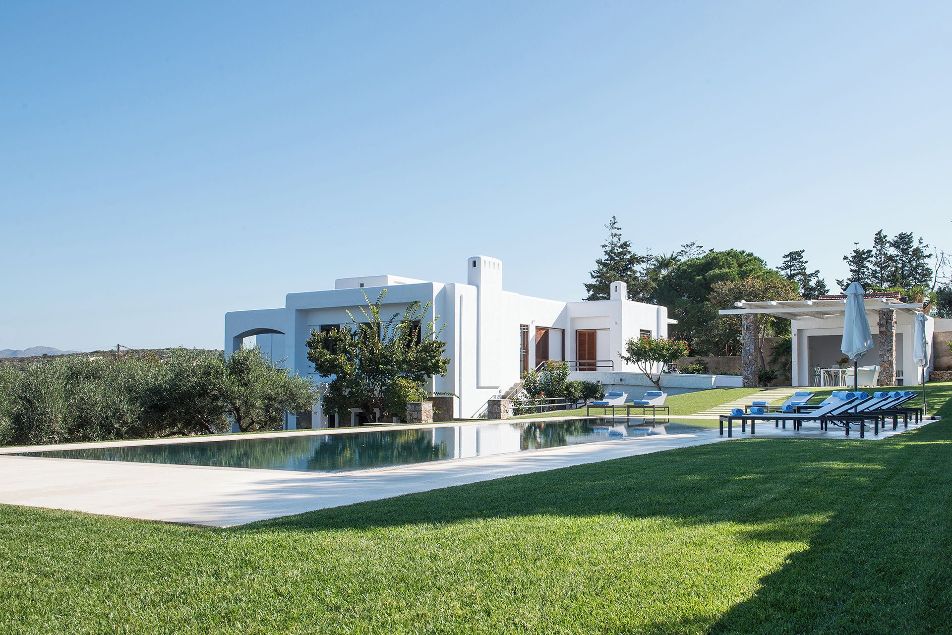 Une villa moderne, de couleur blanche, dotée d  ’une piscine et de chaises longues.