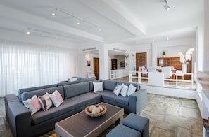 Un vaste espace de vie comprenant un canapé d’angle, une table basse et un coin repas avec un buffet en bois et des meubles blancs.