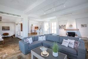 Un salon spacieux avec un grand canapé d’angle gris, une table basse avec des magazines et un chapeau, et une cheminée.