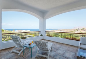 Un balcon avec deux chaises blanches et une table, d’où l’on peut admirer un paysage côtier avec des bâtiments et la mer.