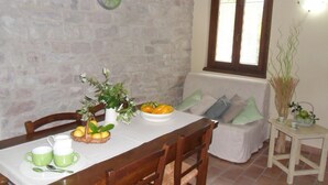 Uma área de refeições com parede de pedra, uma mesa de madeira posta com chávenas verdes, uma tigela de fruta e um vaso com flores.