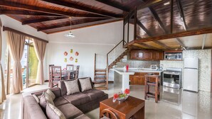 A spacious living area with a sofa, coffee table, and dining table. A kitchen with modern appliances is visible in the background. - Los Arcos dowtown 🌋🌋 (La Fortuna)