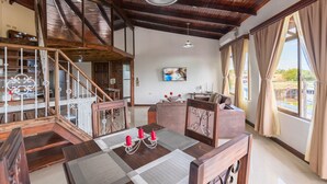 A spacious living room with a high ceiling, wooden beams, a dining area with a table and chairs, a staircase, and large windows offering a view of the outdoors. - Los Arcos dowtown 🌋🌋 (La Fortuna)