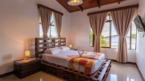 A hotel room with a wooden headboard, a bed with a quilt, two bedside lamps, a wooden nightstand, and a large window with curtains. - Los Arcos dowtown 🌋🌋 (La Fortuna)