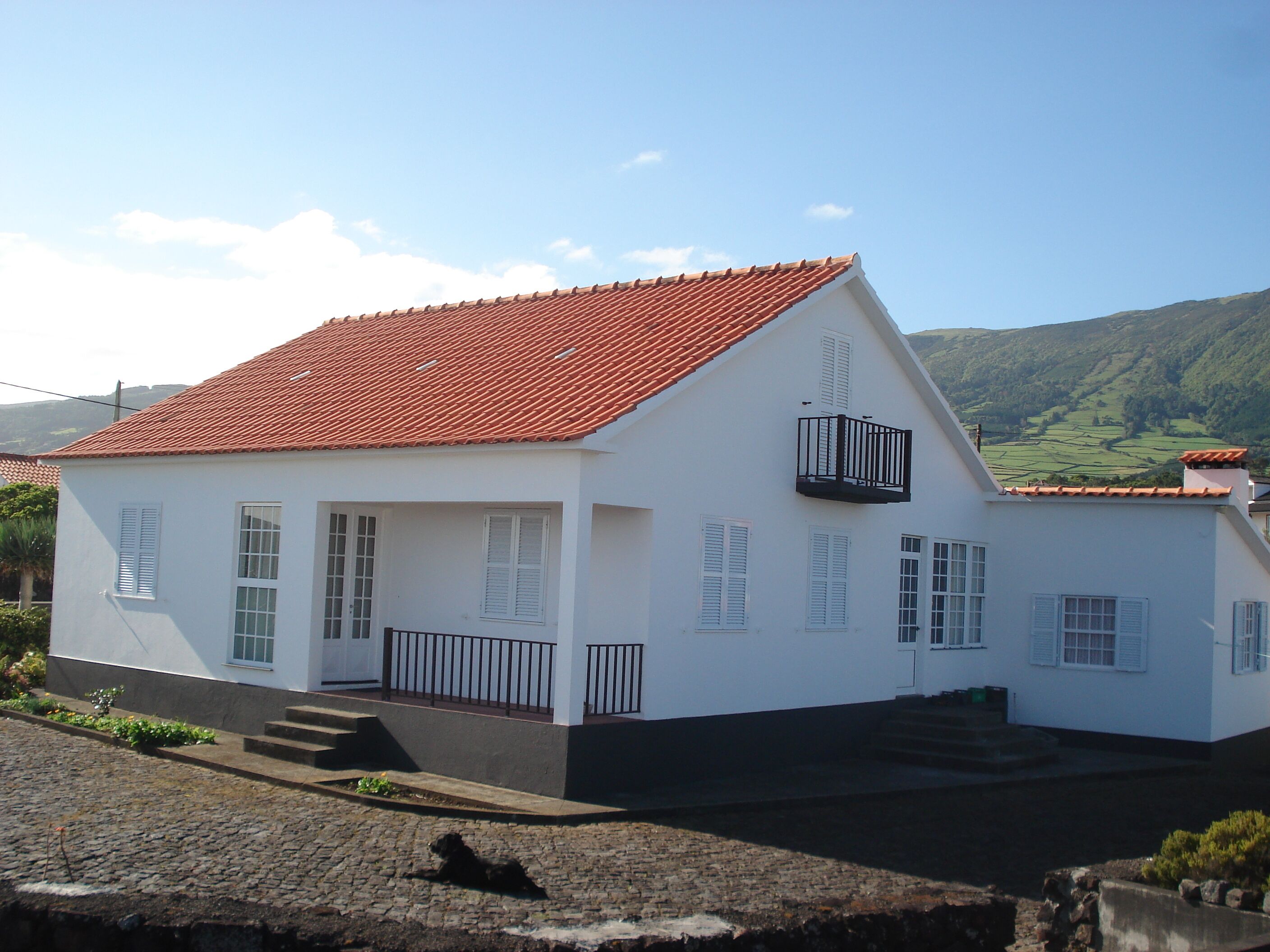 Un bâtiment blanc avec un toit recouvert de tuiles rouges, un balcon et une vue sur des collines verdoyantes en arrière-plan.