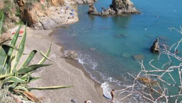 Uma praia costeira com afloramentos rochosos, água azul cristalina e algumas pessoas aproveitando a orla.