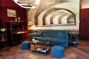 A living room with a blue sofa, a coffee table, and a lamp. There is a brick wall with an arched window and a framed picture on the wall.