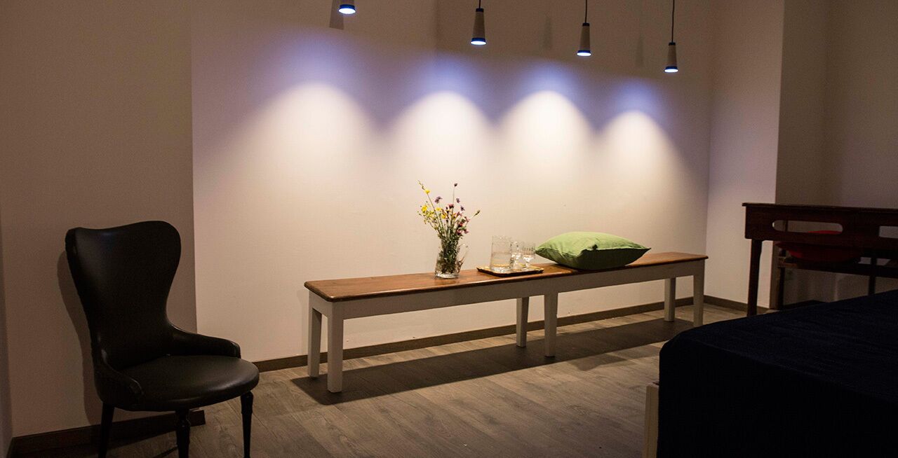 Una habitación con un banco de madera, una pequeña mesa con un florero y un cojín verde, además de una silla negra.