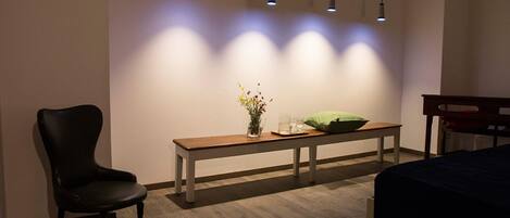 Una habitación con un banco de madera, una pequeña mesa con un florero y un cojín verde, además de una silla negra.