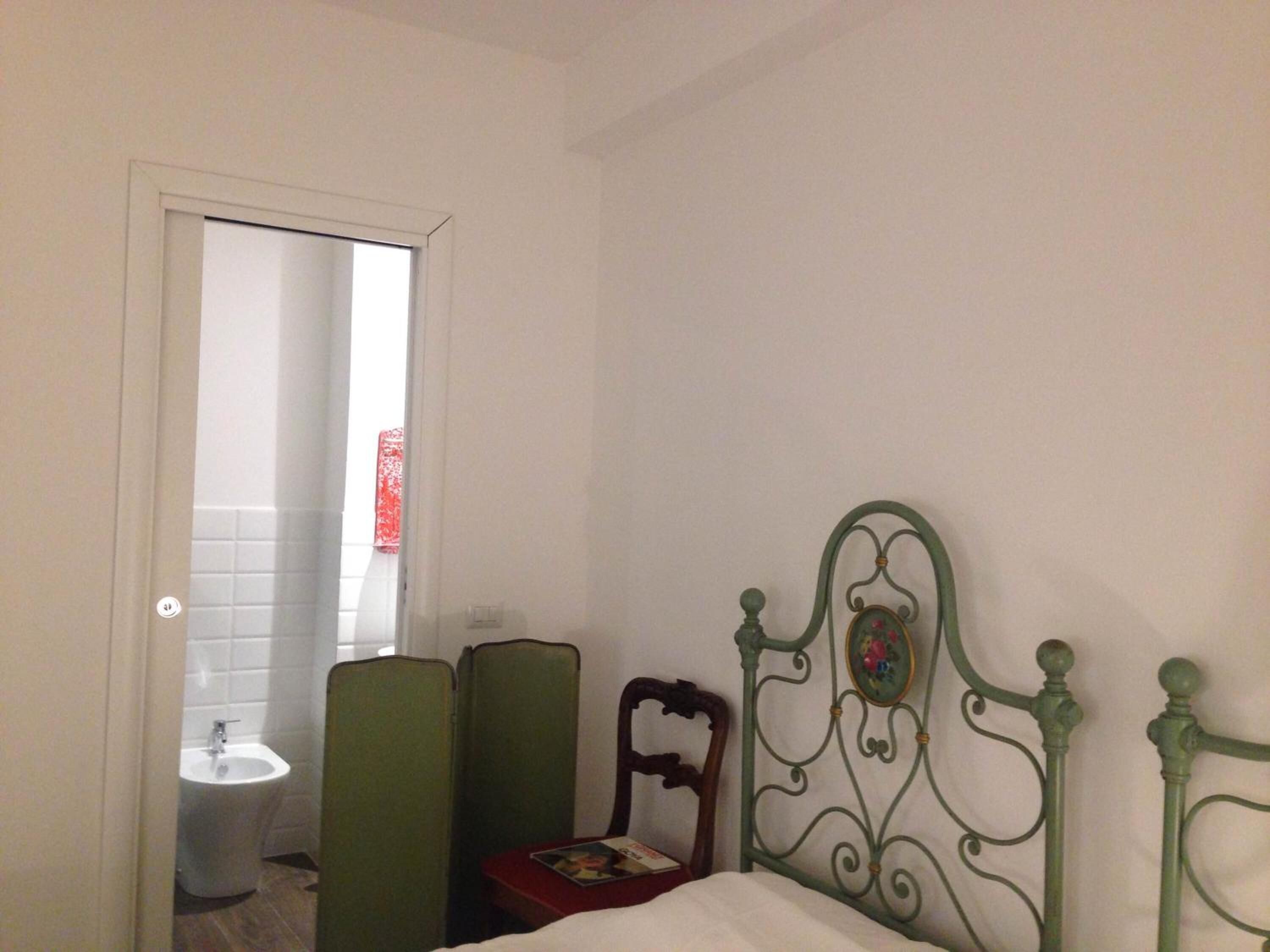 A bedroom with a bed, a wooden chair, a green chair, and a small table with a book.