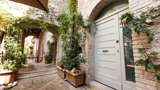 A stone courtyard with arched walkways, potted plants, and a door with a glass panel. - Residenza la Corte Assisi (Assisi)