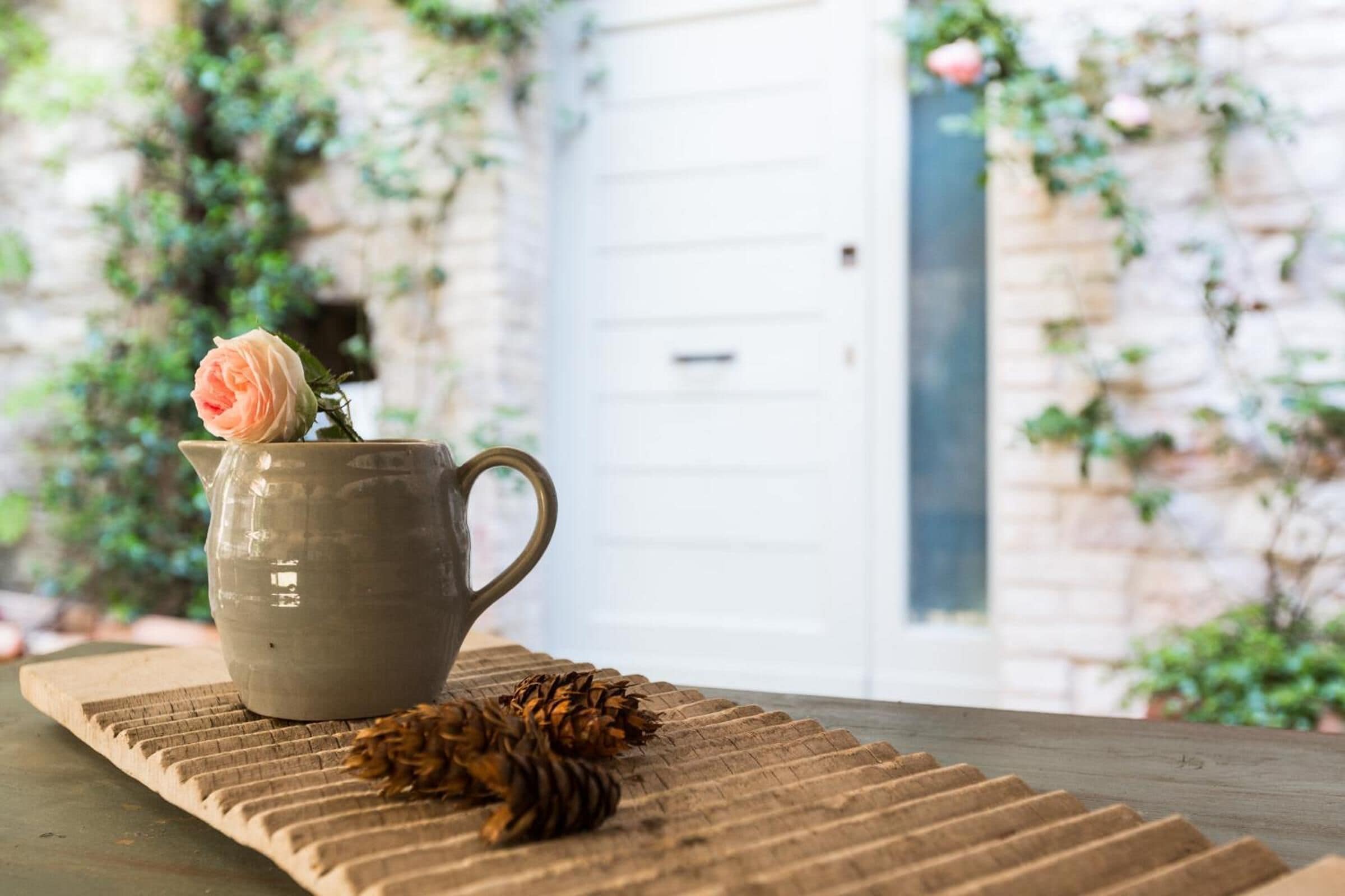 A ceramic pitcher with a single rose on a textured mat, placed on a wooden surface with pine cones.