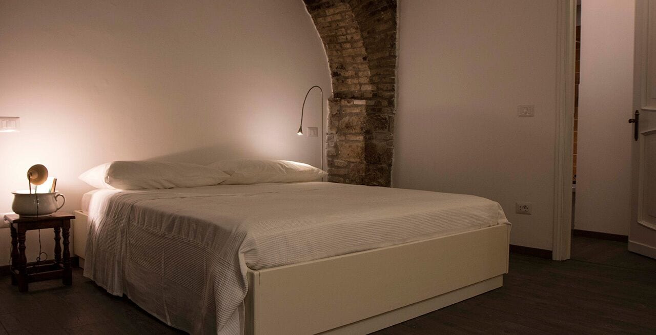 A bedroom with a large bed, a bedside table with a lamp, and a brick wall.
