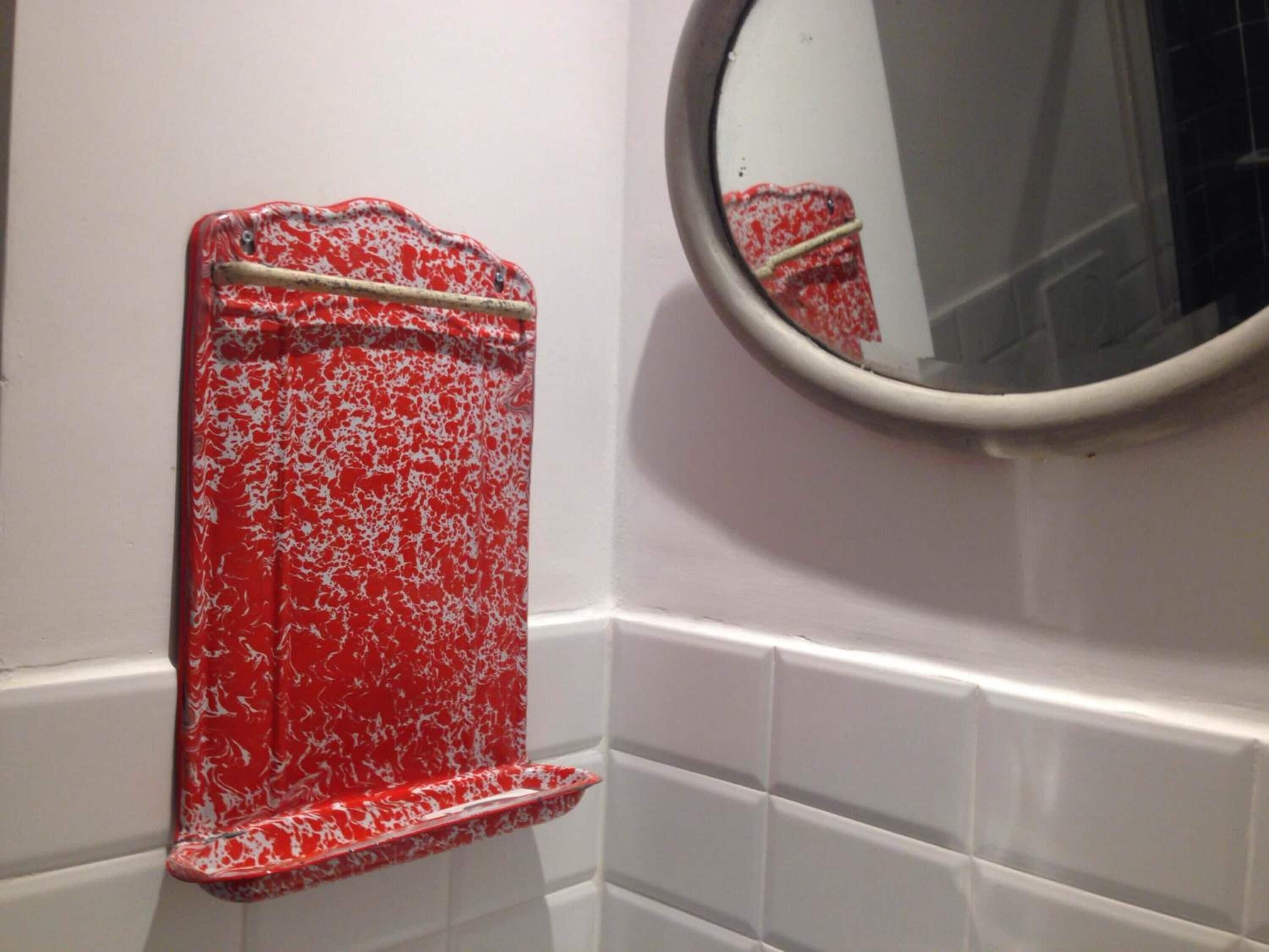 Een badkamer met witte betegelde wanden, een textuurrijke plank in rood en wit, en een ronde spiegel.