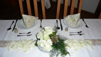 Mesa para dos personas con mantel blanco, platos, cubiertos y un centro de mesa de flores y follaje blanco.