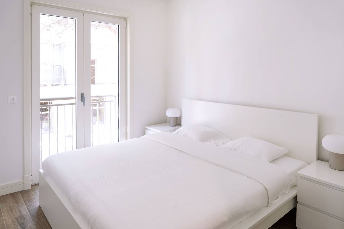 Ein Schlafzimmer mit einem großen Bett, einem Nachttisch, einer Lampe und einer Glastür, die zu einem Balkon im Freien führt.
