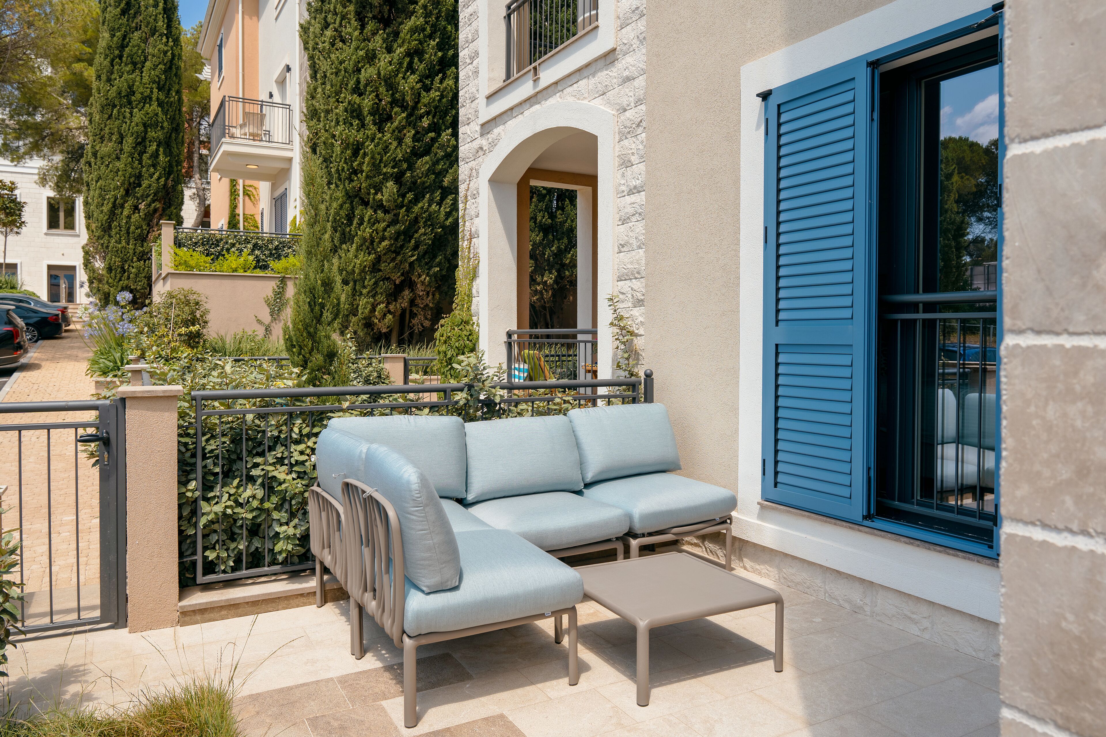 A light blue sofa and a small table on a patio with a stone wall and a blue shuttered window.