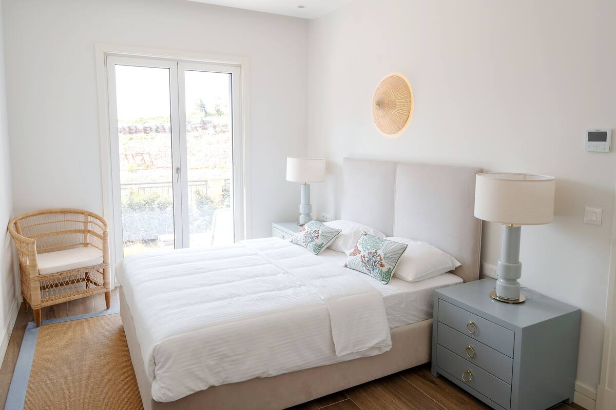 Ein Schlafzimmer mit einem großen Bett, zwei Nachttischen, einem Sessel und Blick durch eine Schiebetür.