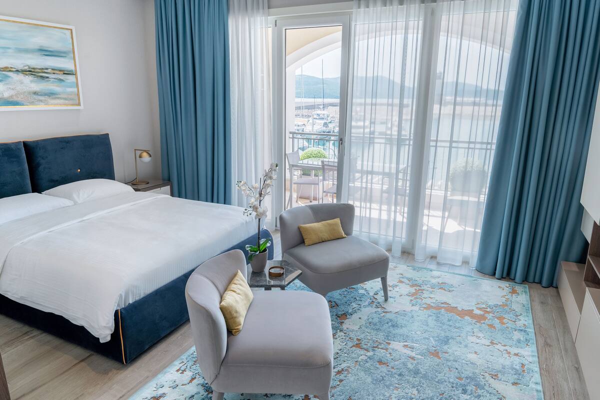 Ein Hotelzimmer mit einem großen Bett, zwei Sesseln, einem kleinen Tisch mit Pflanze und Meerblick durch große Fenster.