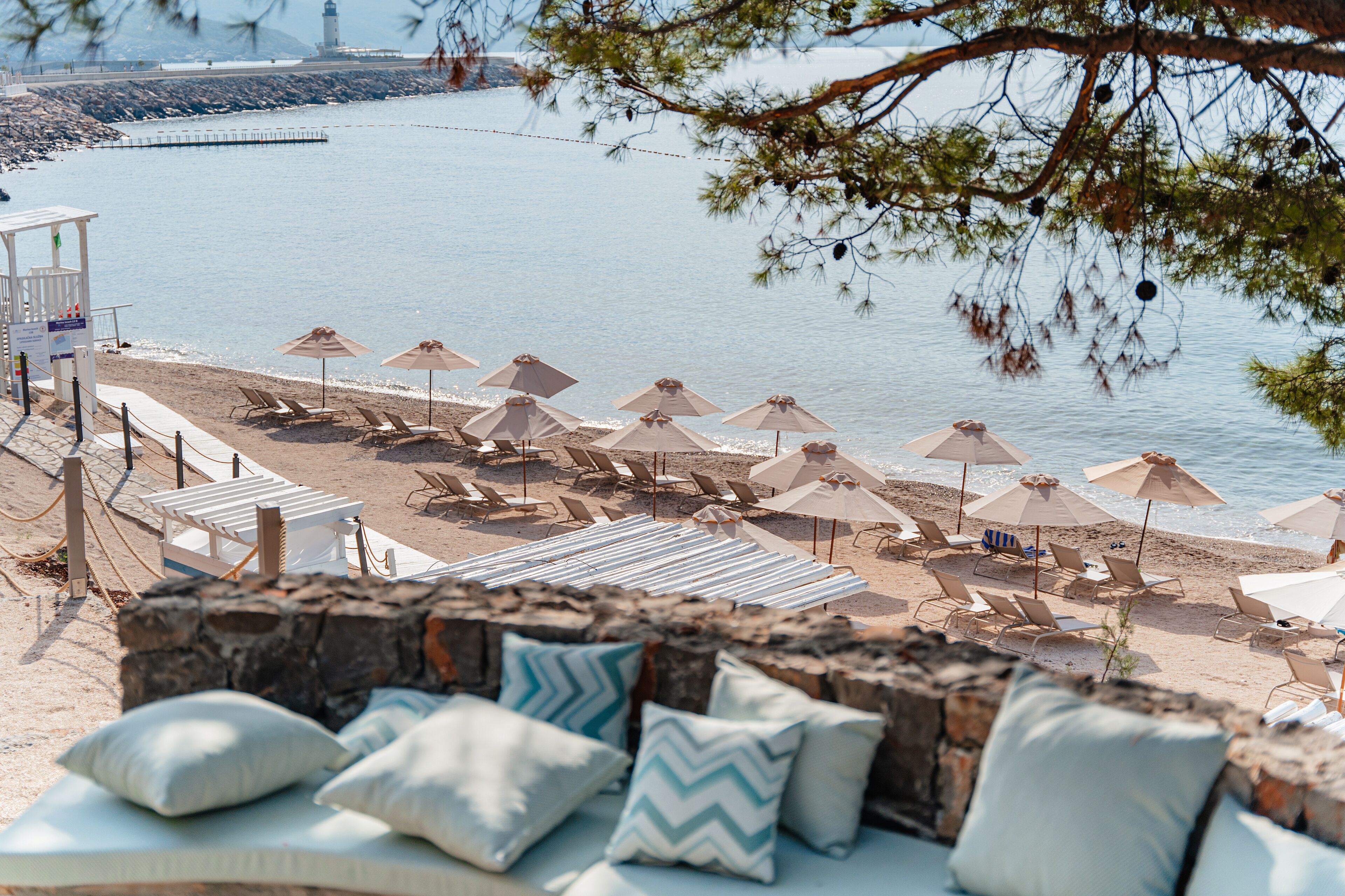 Un área de estar frente a la playa con sillones y sombrillas, con vista al mar tranquilo y un muelle al fondo.