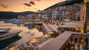Un port esportiu amb iots amarrats, un passeig marítim amb edificis, i una collada de muntanyes al fons.