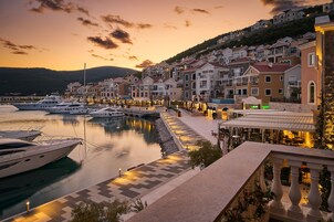 Uma marina com iates atracados, um calçadão à beira-mar com edifícios e uma colina ao fundo.
