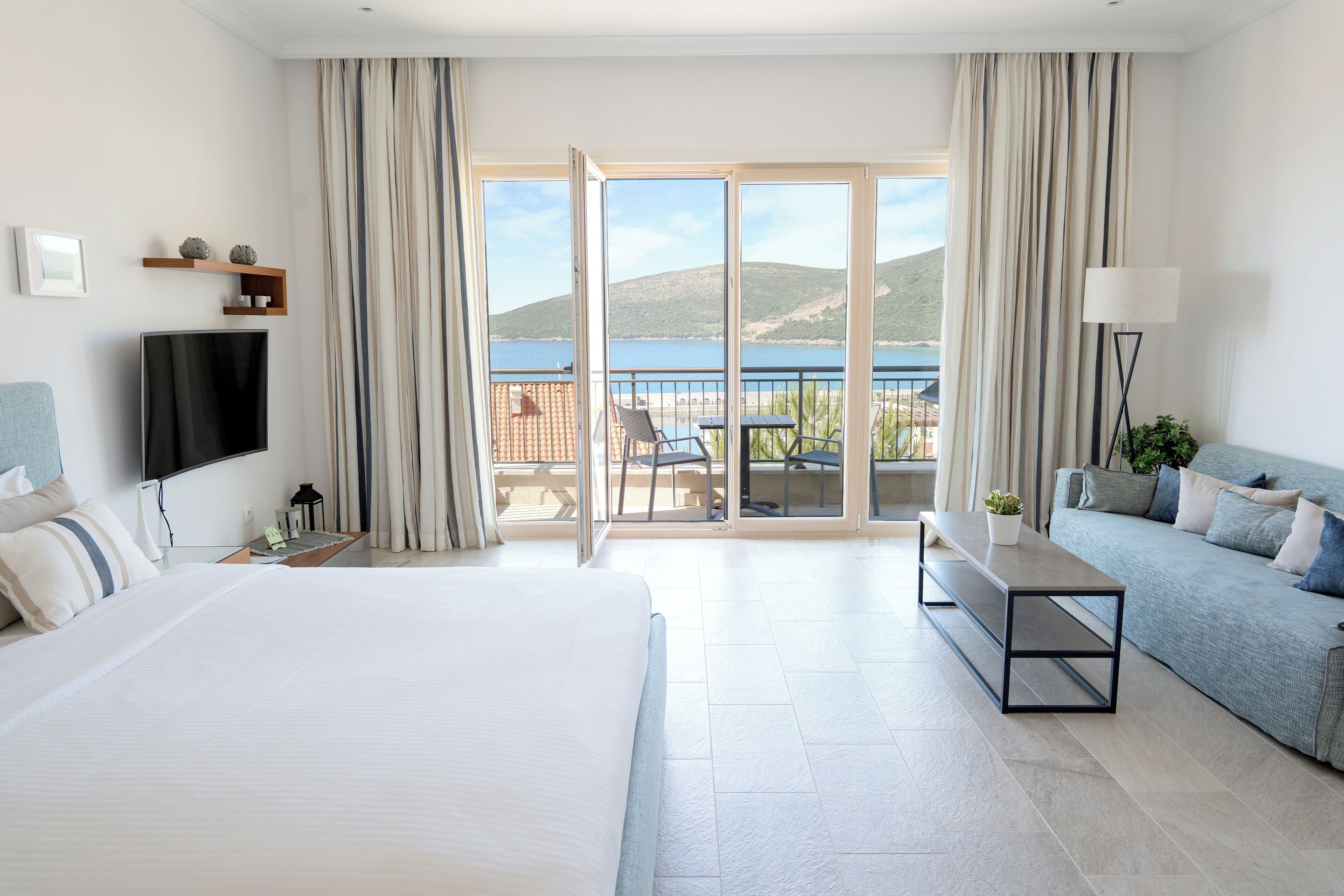 A hotel room with a large bed, a sofa, a TV, and a balcony with a view of the sea.