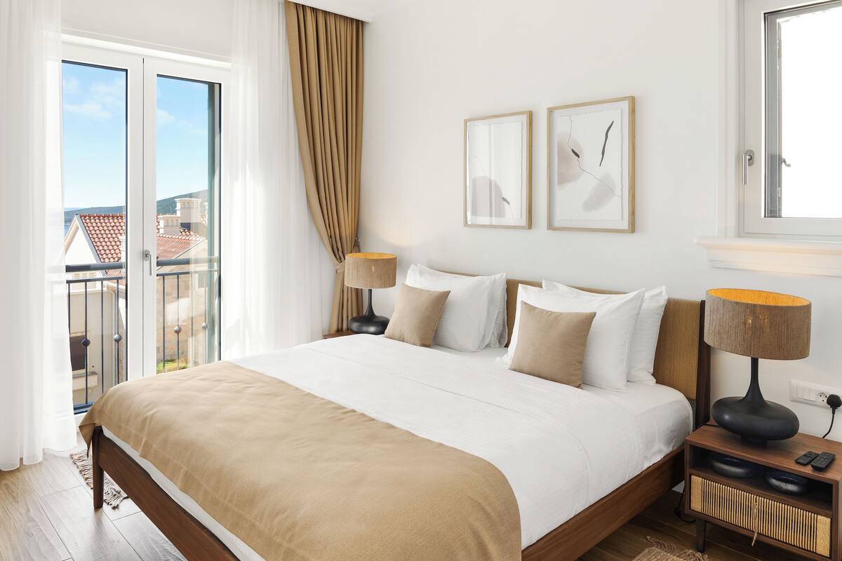 Ein Hotelzimmer mit einem großen Bett, zwei Nachttischlampen, einem Nachttisch und Blick auf die Stadt vom Balkon.