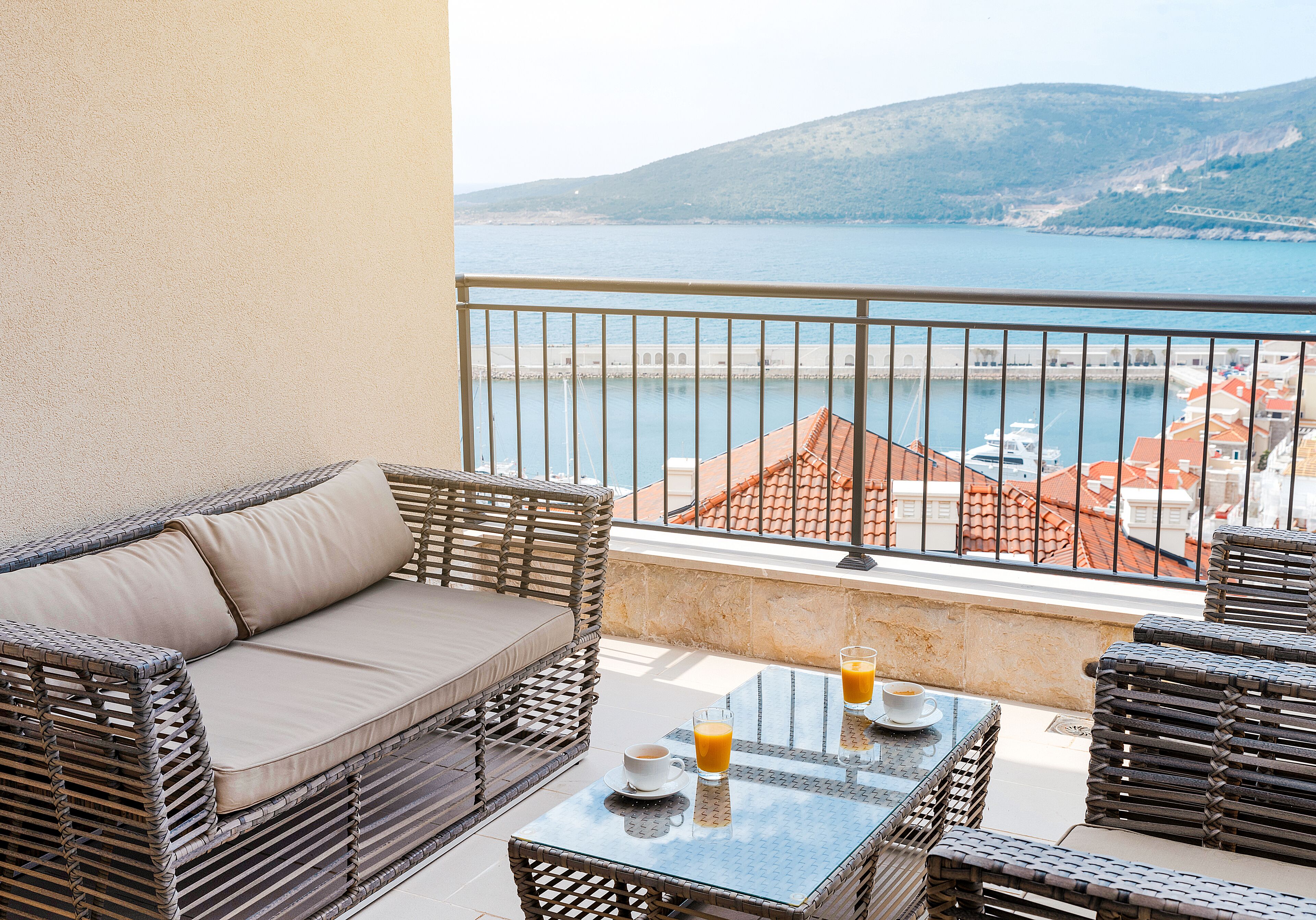 En balkon med fletningsmøbler, et glasbord med drikkevarer og udsigt til havet og bygninger.