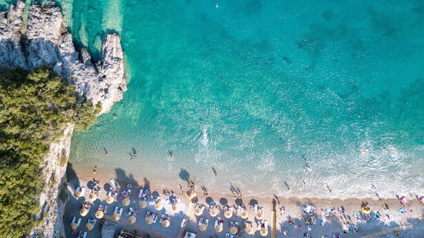 Vista aèria d'una platja amb aigües turqueses cristal·lines, un litoral rocos i una filera de papereres i gandules.