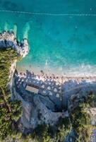 Vista aérea de uma praia com águas turquesa cristalinas, um trecho de costa rochosa e uma fileira de guarda-sóis e espreguiçadeiras.