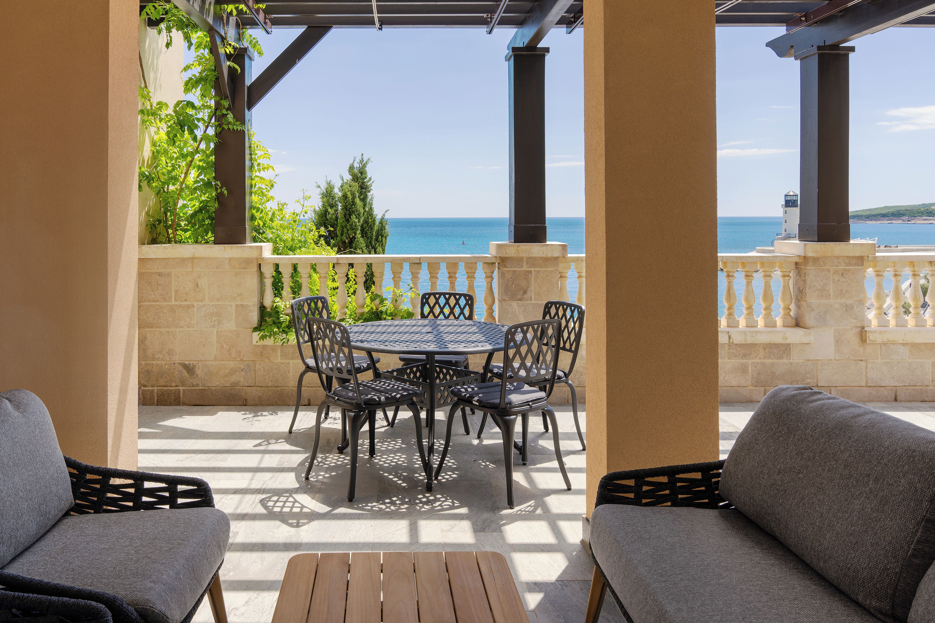 A covered outdoor seating area with a table and chairs, a wooden bench, and a view of the sea.