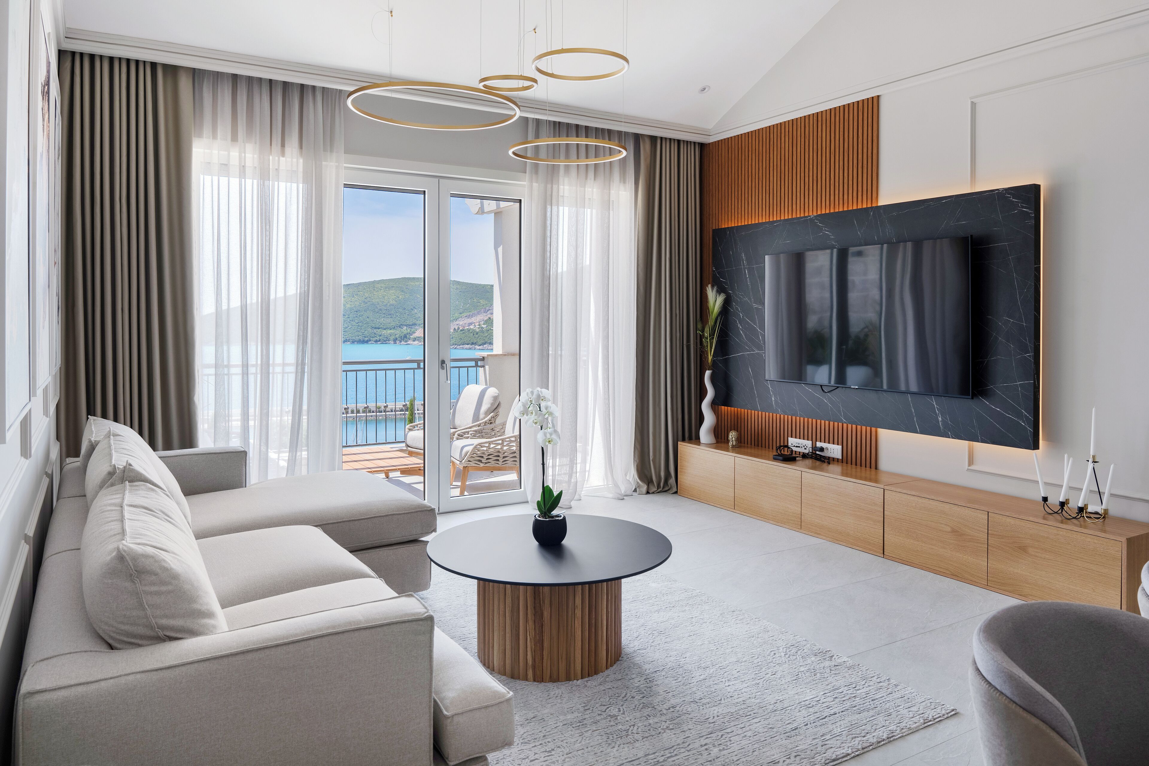 A modern living room with a sofa, a round coffee table, a TV mounted on a wooden panel, and a view of the sea through large windows.