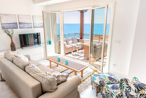 Sala de estar com sofá, mesa de centro e vista para o mar através de grandes portas de vidro.