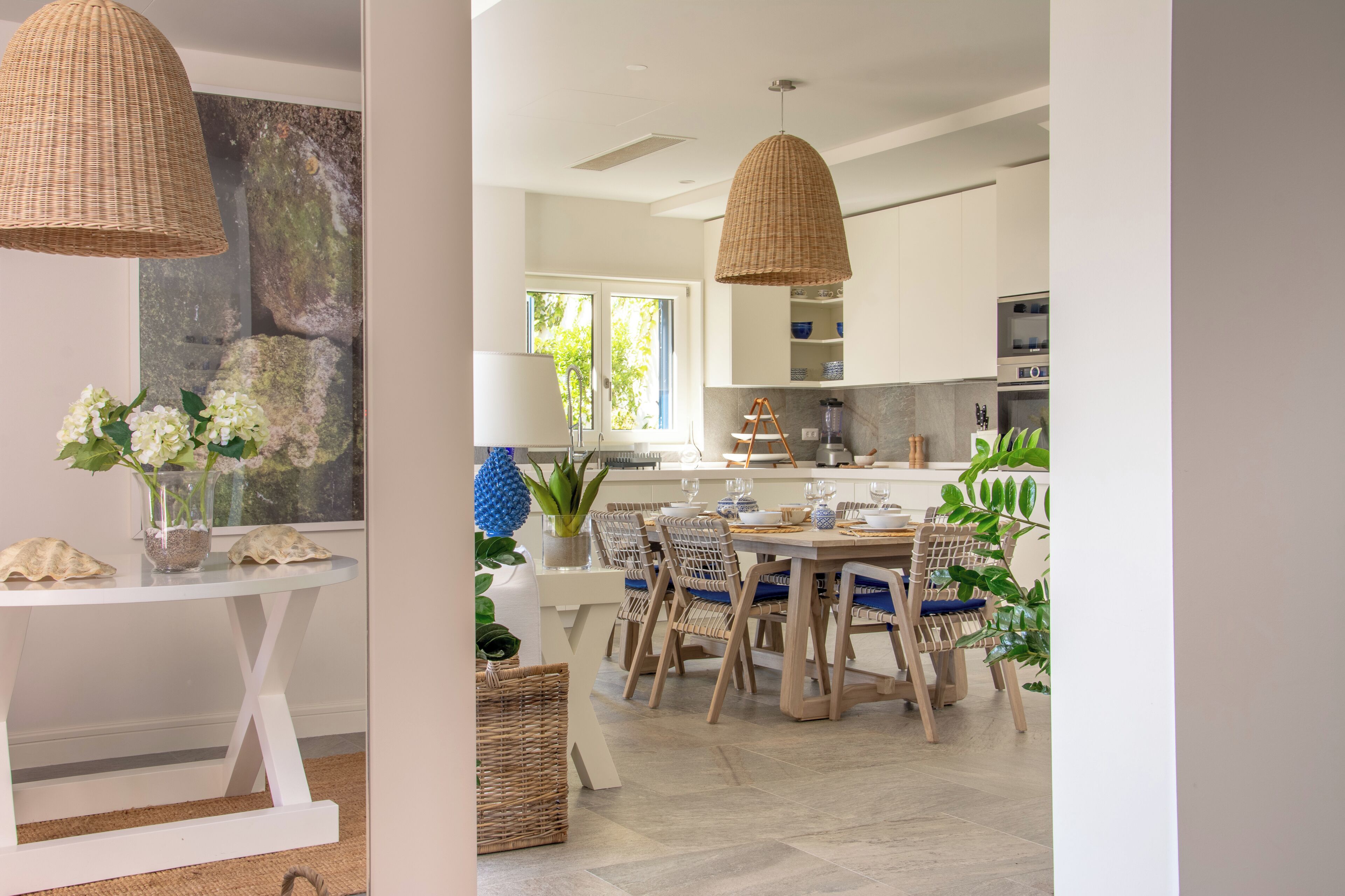 A modern kitchen with white cabinets, a dining area with wooden chairs and a white table, and a hanging wicker light fixture.