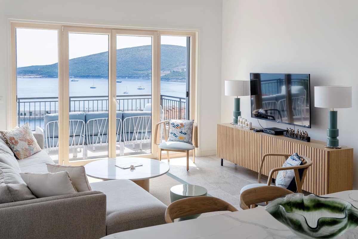 Ein modernes Wohnzimmer mit großem Fernseher, bequemen Sitzmöglichkeiten und einem Balkon mit Meerblick.