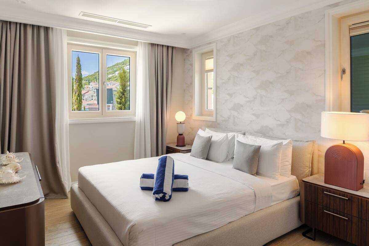 Ein Hotelzimmer mit einem großen Bett, einem Nachttisch mit Lampe, Blick ins Freie durch ein Fenster und einem Holznachtkästchen.