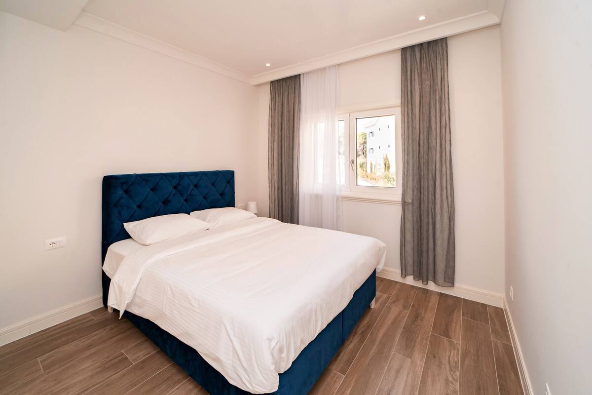 Ein Schlafzimmer mit blauem Kopfteil, weißer Bettwäsche und einem Fenster mit grauen Vorhängen.