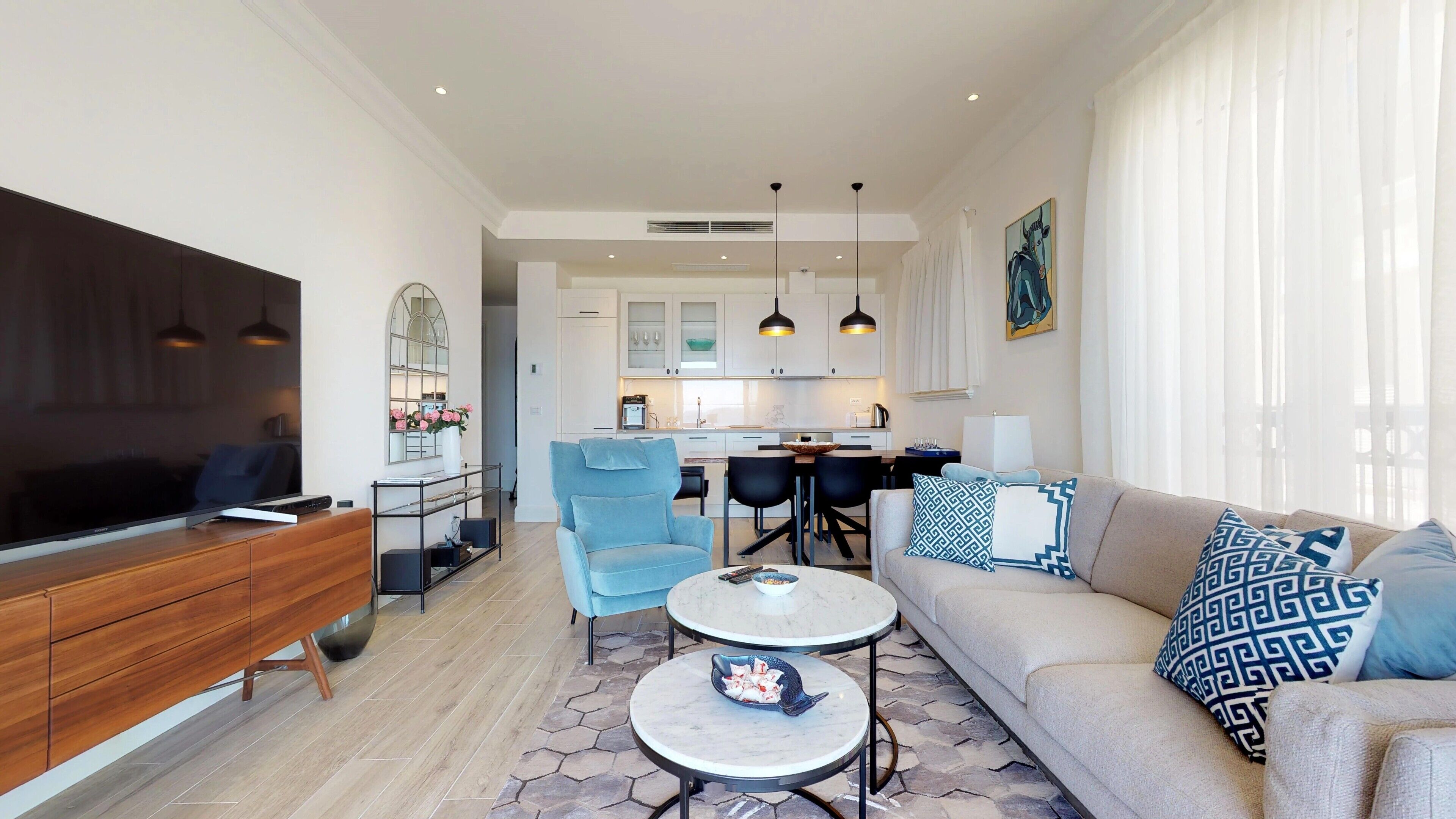Una sala moderna con televisor, sofá y mesa de centro. Se ve una cocina al fondo con armarios blancos y lámparas colgantes.
