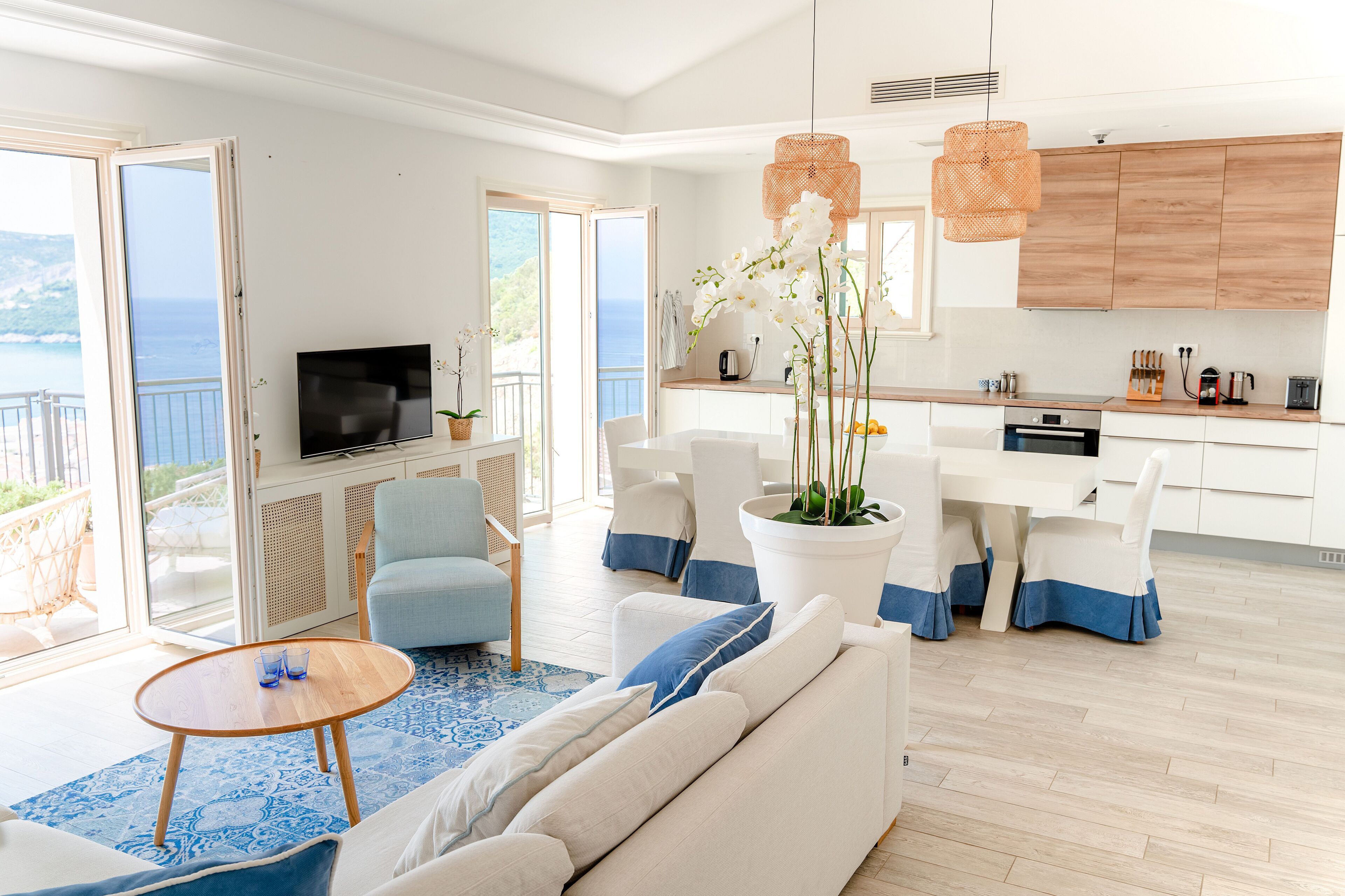 A modern living room with a sofa, a coffee table, and a TV. There is a balcony with a view of the sea.