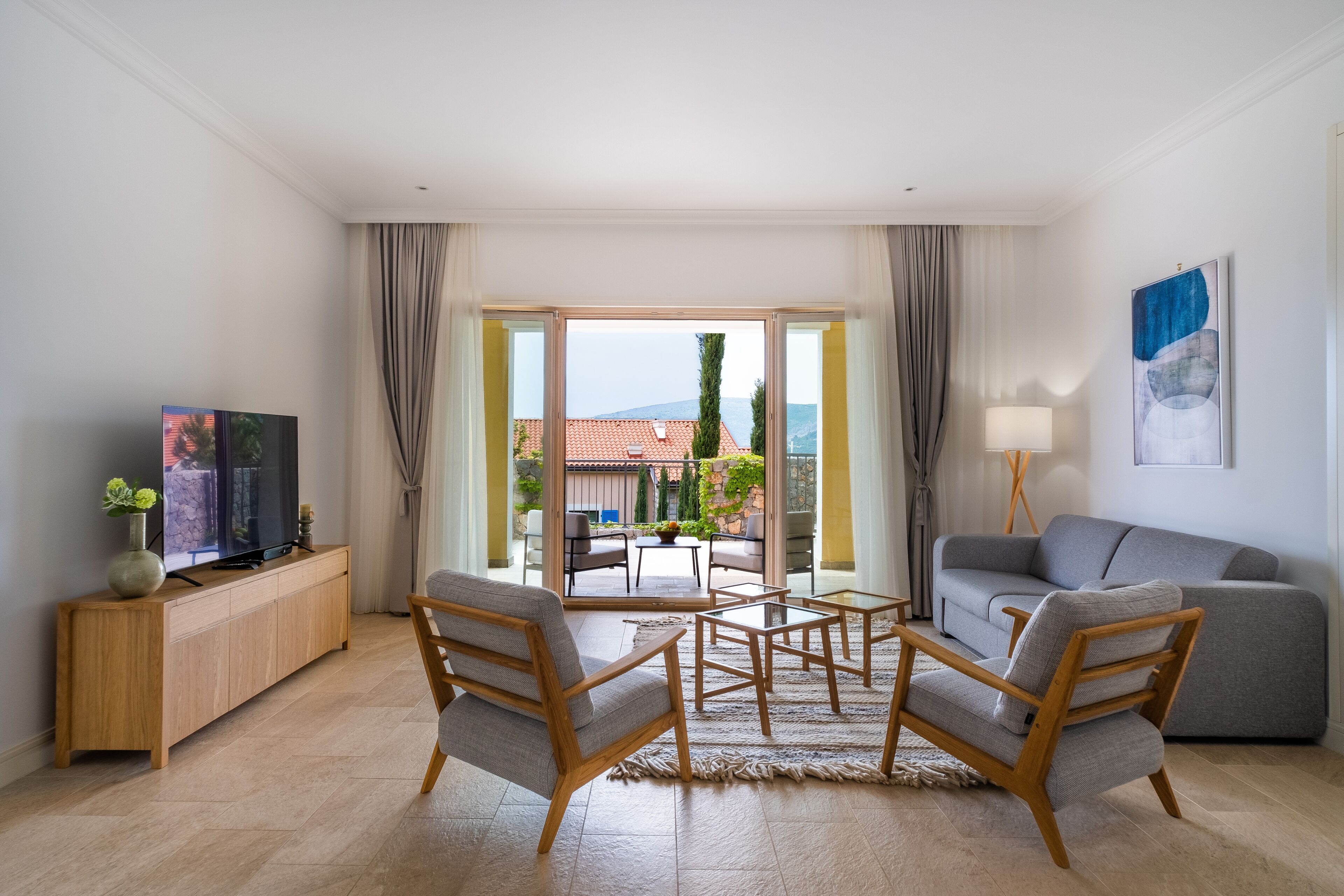 Un salon moderne avec un téléviseur, un canapé, des fauteuils et une vue sur l’extérieur, accessible par une porte vitrée.