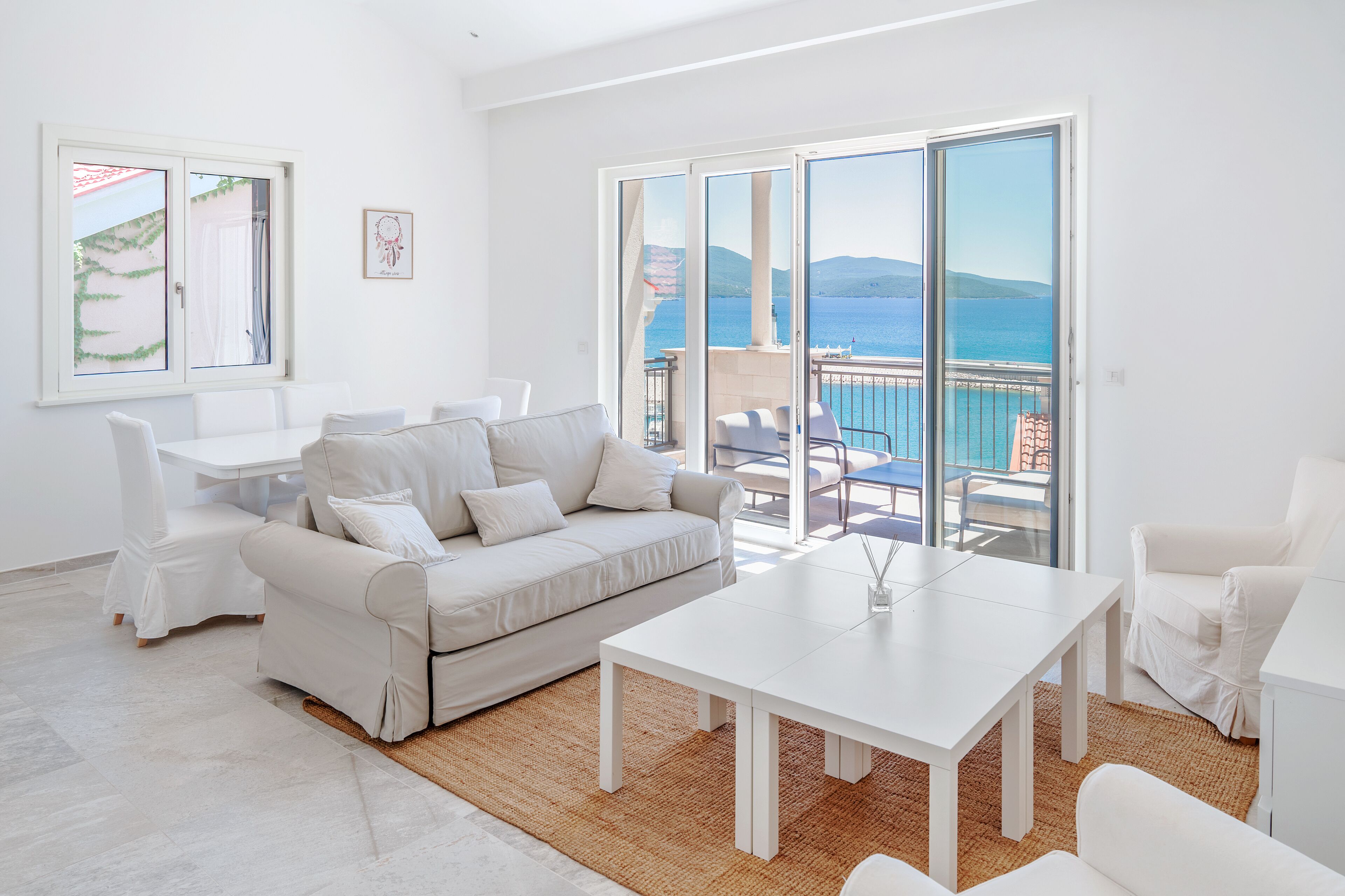 A modern living room with a white sofa, a coffee table, and a dining table. There is a large sliding glass door leading to a balcony with a view of the sea and mountains.