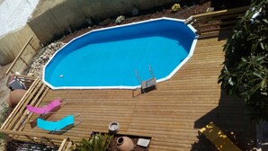 Een buitenzwembad met een houten terras, twee ligbedden en een kleine tuin met planten en een decoratieve pot.