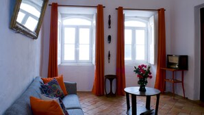 Een woonkamer met een bank, oranje gordijnen, een televisie op een dressoir en een vaas met bloemen.