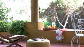 Une terrasse avec un fauteuil de relaxation en bois, une chaise en métal blanc et un pouf rond.