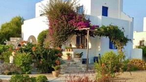 Un bâtiment blanc avec une porte bleue et un porche couvert orné de plantes. Des marches mènent au porche, et un mur de pierre se trouve à l’arrière-plan.