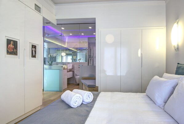 Una habitación de hotel moderna con una cama bien hecha, un baño con cabina de ducha de vidrio y una pequeña zona de estar.