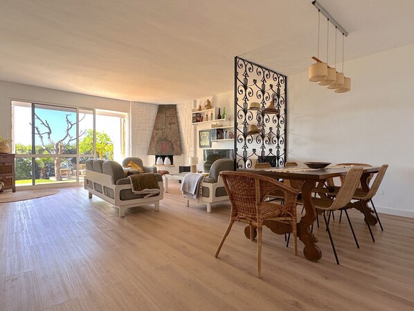 A spacious living room with a fireplace, large windows, and a dining area with wicker chairs.