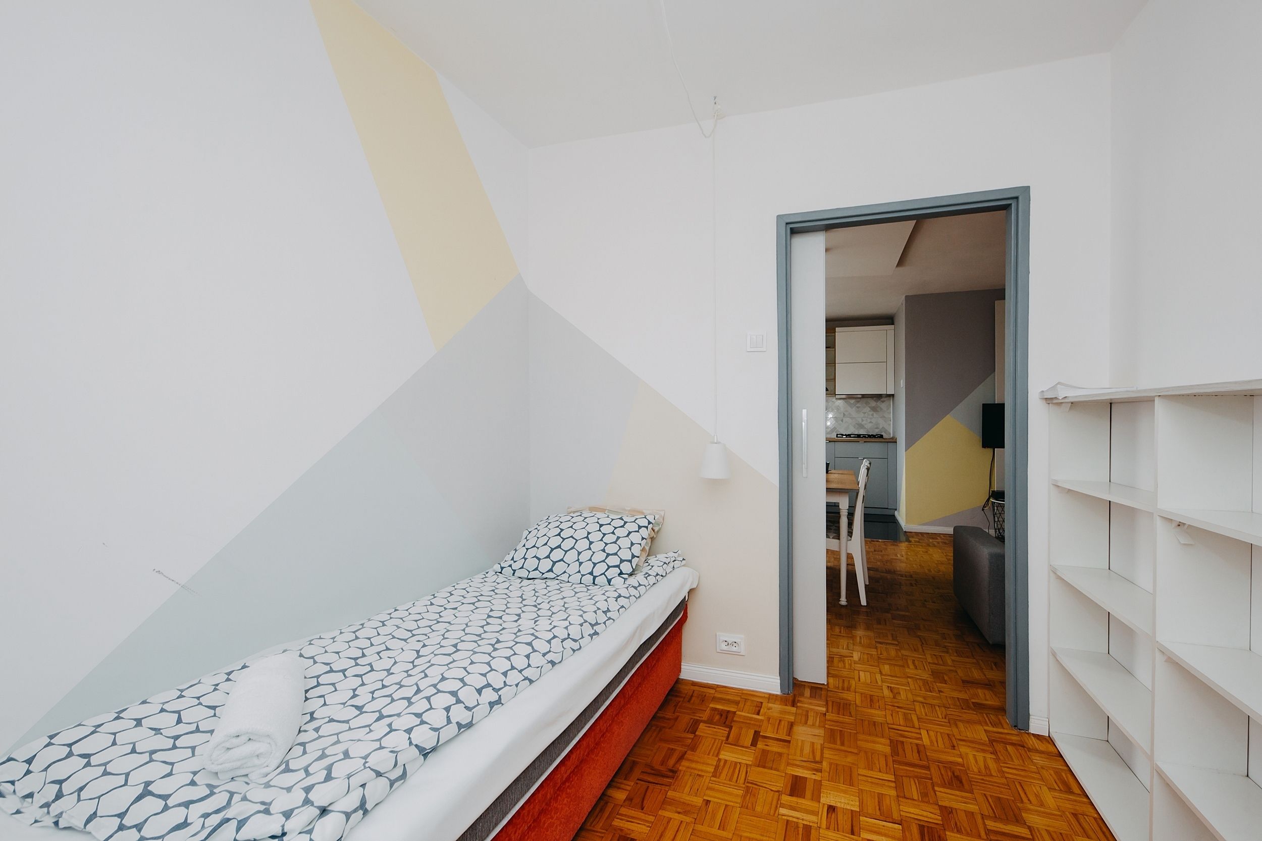 Een kleine, compacte kamer met een enkel bed, een houten vloer en een doorgang naar een keukenhoek.
