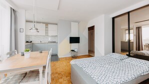 Una habitación de hotel moderna con una mesa de comedor de madera, una cama con ropa de cama estampada, un televisor montado en la pared y una cocina visible a través de una puerta abierta.